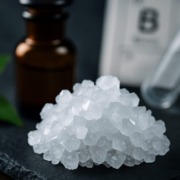 Makroaufnahme von weißen Borax-Kristallen vor einem Apothekerfläschchen und dem Elementsymbol für Bor – Symbolbild für naturheilkundliche Anwendung