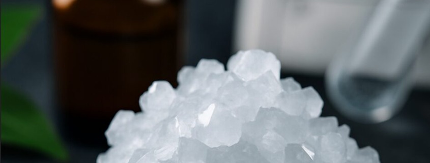Makroaufnahme von weißen Borax-Kristallen vor einem Apothekerfläschchen und dem Elementsymbol für Bor – Symbolbild für naturheilkundliche Anwendung